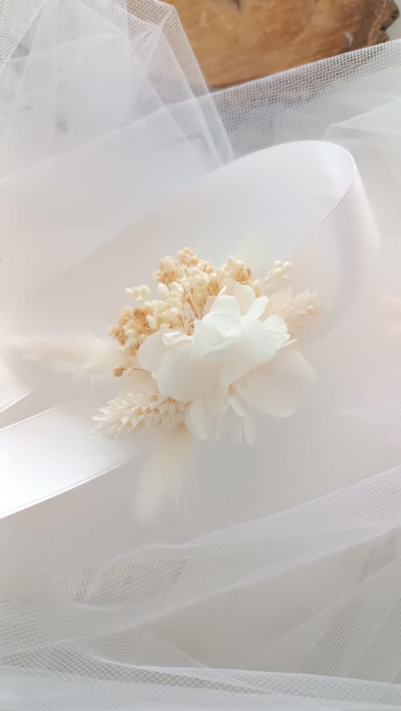 May include: A white flower bouquet with dried flowers and a white ribbon. The bouquet is on a white tulle background.