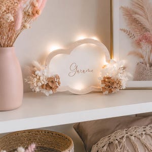 Può includere: Un cartello di legno a forma di nuvola con il nome "Garance" illuminato da piccole luci. Il cartello è decorato con fiori secchi e si trova su una mensola bianca con un vaso di fiori rosa e un cestino intrecciato.