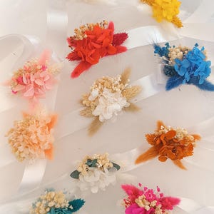 May include: Assortment of dried flower wrist corsages in various colors, including pink, white, red, orange, yellow, blue, and teal. Each corsage is tied with a white satin ribbon.