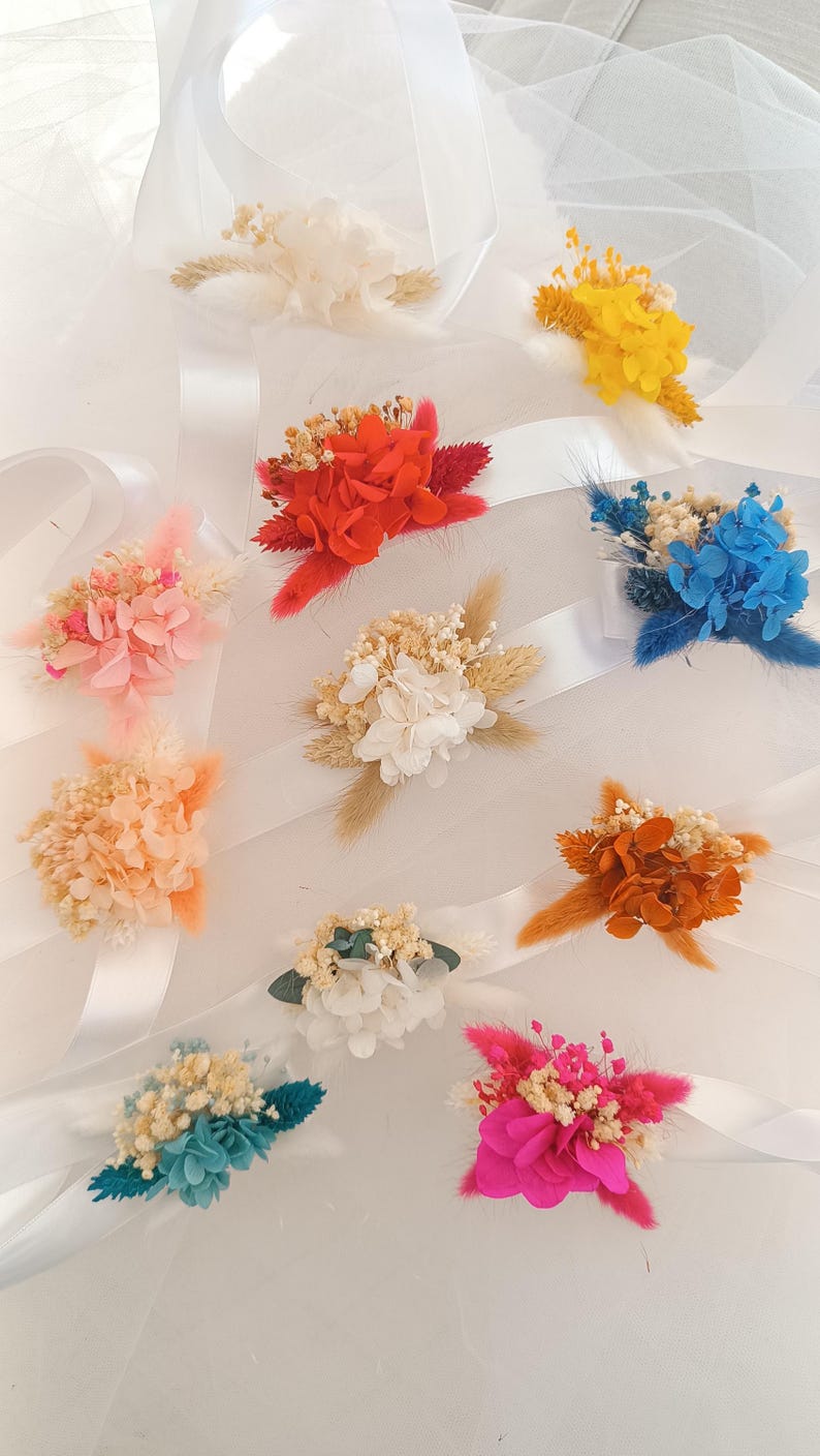 May include: Assortment of dried flower wrist corsages in various colors, including pink, white, red, orange, yellow, blue, and teal. Each corsage is tied with a white satin ribbon.