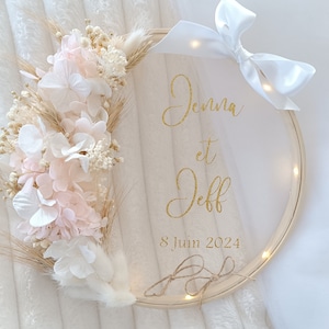 Może przedstawiać: Biało-beżowy wieniec kwiatowy z białą kokardą jest umieszczony na drewnianym obręczy do haftu. Obręcz ma biały tiulowy podkład z złotym napisem "Jenna et Jeff 8 Juin 2024".