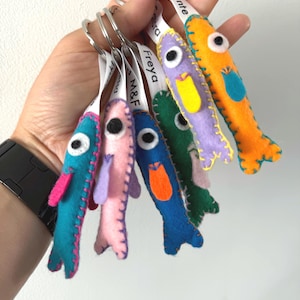 Puede incluir: Un conjunto de seis llaveros de fieltro con forma de pez en colores vibrantes como turquesa, rosa, azul, verde, morado y naranja. Cada pez presenta un ojo blanco con pupila negra, una aleta de fieltro en contraste y una cinta blanca.