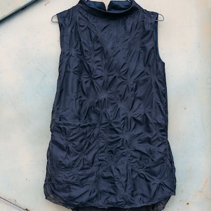 Può includere: Top senza maniche blu navy con collo alto e motivo trapuntato strutturato. L'indumento è appeso a una gruccia di metallo su uno sfondo azzurro chiaro usurato. L'orlo inferiore presenta un design asimmetrico e a strati.