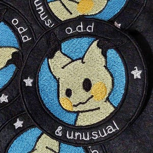 May include: Three embroidered patches featuring a cartoon character with yellow ears and a blue background. The patches are circular and have black borders with white text that reads "odd & unusual".