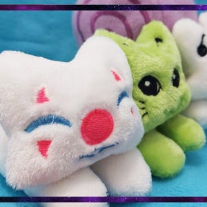 May include: A close-up of three plush toys in a row. The first is white with pink and blue accents, the second is green, and the third is white. The toys are on a blue surface.