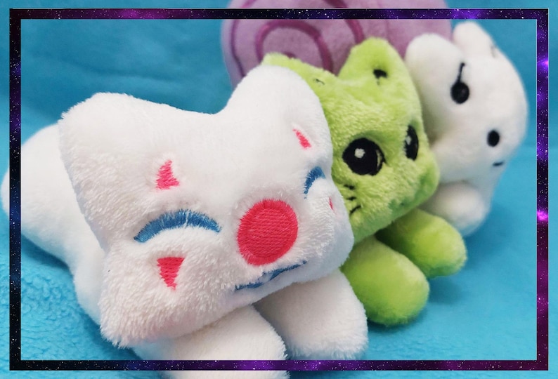 May include: A close-up of three plush toys in a row. The first is white with pink and blue accents, the second is green, and the third is white. The toys are on a blue surface.
