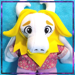 Puede incluir: Un peluche con una cara blanca, ojos grandes y un hocico largo. Tiene pelo amarillo y un cuello amarillo, y viste una camisa y pantalones cortos de flores rosas. El juguete tiene cuernos blancos.