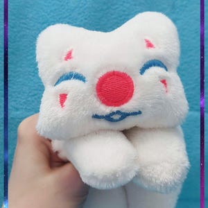 May include: A white plush toy with a smiling face, featuring a red circle nose, blue eyebrows, and small pink accents. The toy is held against a blue background with a purple and blue border.