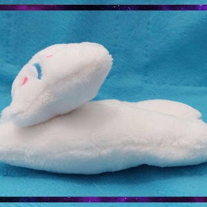 May include: A white plush cloud-shaped pillow with a small face embroidered with blue and pink details. The pillow is on a blue background and framed with a purple and black border.