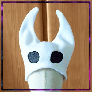 May include: White plush hat with two upward-curving horns and two large, black, embroidered eye-like shapes. The hat is designed to resemble a character from a video game. The hat is displayed on a mannequin head.