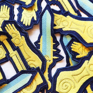Pode incluir: Uma coleção de patches bordados para passar a ferro com vários designs em dourado e azul. Os patches estão sobre um fundo branco.