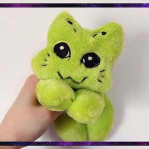以下が含まれることがあります： 黒い目と笑顔の口を持つ、猫の形をしたライムグリーンのぬいぐるみ。おもちゃは柔らかくふわふわで、小さな腕と尖った耳のある頭があります。背景は白で、紫と青のボーダーがあります。