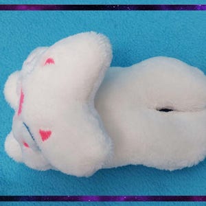 May include: A white plush toy in the shape of a cloud, with pink heart-shaped accents. The toy is lying on a blue surface, framed by a purple and black border. The plush toy is soft and fluffy.