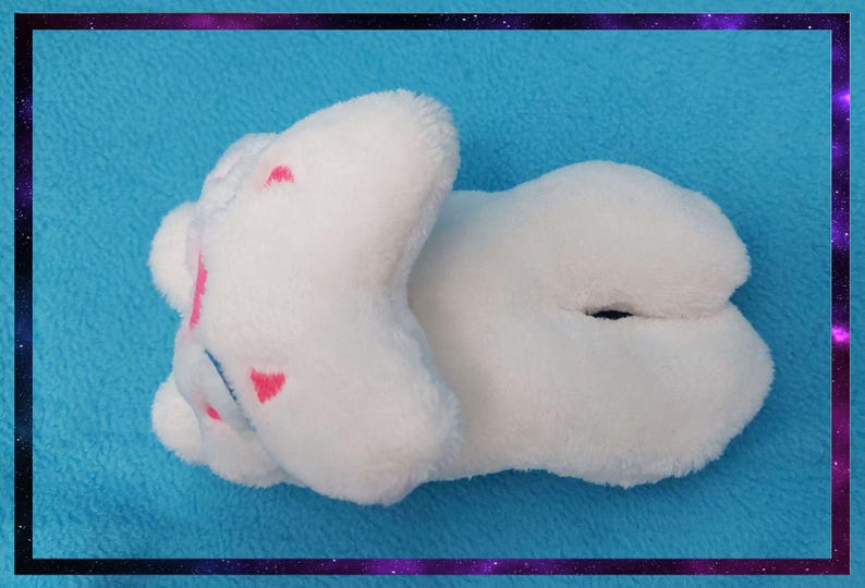 May include: A white plush toy in the shape of a cloud, with pink heart-shaped accents. The toy is lying on a blue surface, framed by a purple and black border. The plush toy is soft and fluffy.