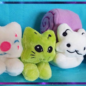 May include: A collection of plush cat toys in various colors. The toys include white, green, and blue cats, and a purple snail. The white cat has pink and blue facial details. The green cat has black eyes and whiskers.