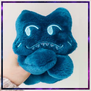 May include: A plush, teal-colored creature toy with embroidered facial features, including eyes and a toothy grin. The soft toy is held in a person's hand, showcasing its fluffy texture and whimsical design. The toy is framed by a purple and black border.