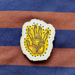 Puede incluir: Una mano amarilla con líneas y puntos negros sobre un fondo blanco. La mano está sobre un círculo de madera.