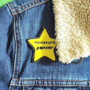 May include: A yellow star-shaped patch sewn onto a blue denim jacket. The patch has black stitching around the edges and the words "miniature painter" written in black text.
