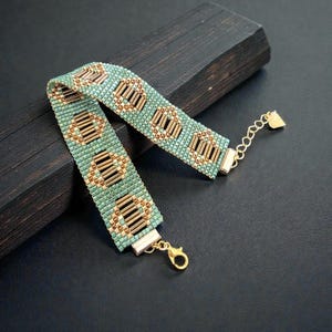 Op de afbeelding: Een kralenarmband met een turquoise en gouden geometrisch patroon. De armband heeft kleine turquoise kralen en goudkleurige buiskralen in een herhalend ontwerp. Het heeft een goudkleurige sluiting en ketting voor verstelbare maat.