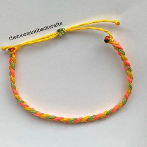 May include: A braided bracelet featuring yellow, orange, and green threads. The bracelet has an adjustable yellow cord with a green knot. The text "themoonandbackcrafts" is visible in the image.