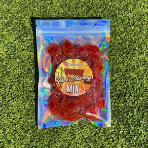 Puede incluir: Una pequeña bolsa de plástico transparente llena de trozos de caramelo rojo, naranja y amarillo. La bolsa tiene un acabado holográfico y una pegatina que dice "Yari's Chamoy MIX".