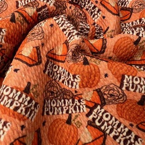 May include: Close-up of a textured orange fabric with a repeating pattern of pumpkins, floral designs, and the text "Mommas Pumpkin" in white and brown. The fabric is folded, showing depth and detail.