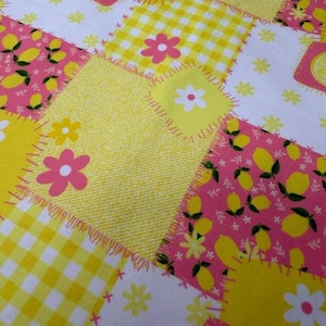 May include: A patchwork fabric with a cheerful design. The fabric features squares of yellow, white, and pink, adorned with lemons, flowers, and gingham patterns. The edges of the squares are stitched with orange thread.