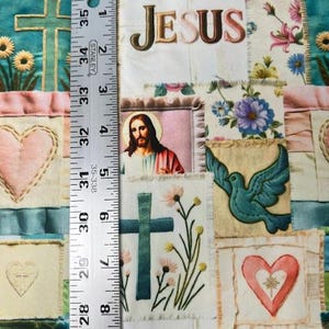 May include: A patchwork fabric with religious imagery. The design includes embroidered crosses, hearts, flowers, and the word "JESUS". The fabric has a variety of colors, including teal, pink, and white. The ruler shows a measurement of 35 inches.