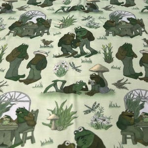 May include: A light green fabric featuring a whimsical pattern of anthropomorphic frogs in various poses. The frogs are depicted in green and brown attire, engaged in activities like reading, socializing, and relaxing in a garden setting with mushrooms, flowers, and dragonflies.