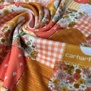 May include: A patchwork quilt with a variety of floral, gingham, and solid fabric squares in shades of orange, peach, yellow, and white. The quilt has a textured appearance with visible stitching and the word "carhartt" printed on one of the squares.