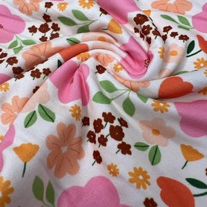 May include: White fabric with a vibrant floral pattern. Large pink flowers are interspersed with smaller orange and yellow blooms, brown clusters, and green leaves. The fabric is soft and has a slight texture, perfect for apparel or home decor.
