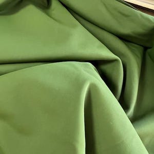 May include: Close-up of olive green fabric with soft folds and a smooth texture. The fabric appears to be a solid color, with variations in tone due to the folds. The material is likely used for clothing or home decor.