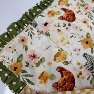 May include: A white fabric with a floral and chicken pattern. The design features brown, yellow, and pink flowers, with images of chickens. The edge is trimmed with a green polka dot ruffle. A yellow and white checkered pattern is visible at the bottom.