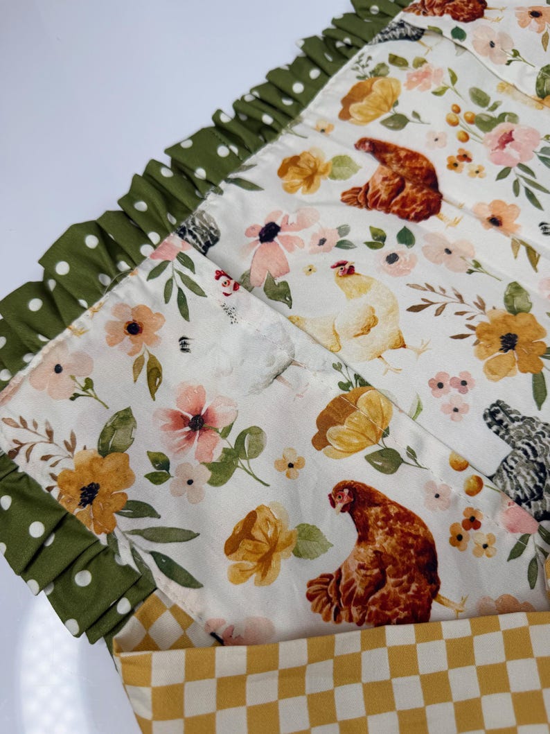 May include: A white fabric with a floral and chicken pattern. The design features brown, yellow, and pink flowers, with images of chickens. The edge is trimmed with a green polka dot ruffle. A yellow and white checkered pattern is visible at the bottom.