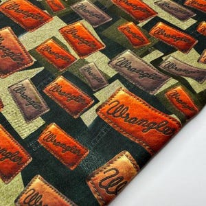 May include: Close-up of a fabric with a repeating pattern of rectangular patches. The patches are in shades of brown and orange, with the word "Wrangler" in a script font. The background has a geometric design in green and black.
