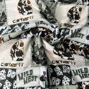 Op de afbeelding: Witte stof met een patchwork-ontwerp met een bruine koehuidprint, speelkaarten en de tekst "Wild West" en "Carhartt".