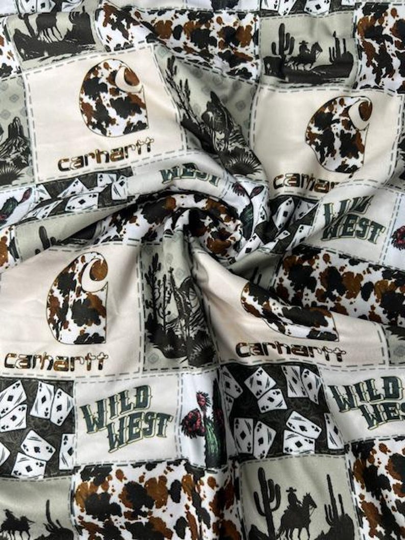 Op de afbeelding: Witte stof met een patchwork-ontwerp met een bruine koehuidprint, speelkaarten en de tekst "Wild West" en "Carhartt".