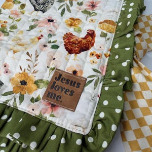 May include: A quilted, rectangular fabric with a white background featuring floral and chicken illustrations. The border is green with white polka dots, and a ruffle edge. A brown tag reads "Jesus loves me."