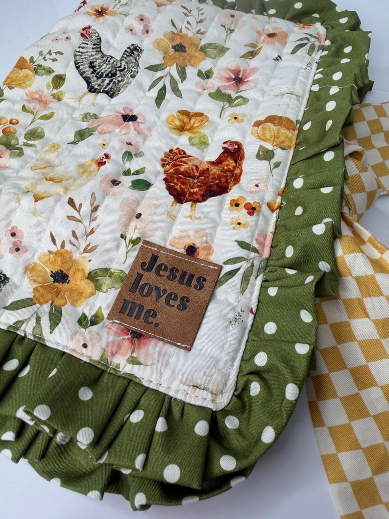 May include: A quilted, rectangular fabric with a white background featuring floral and chicken illustrations. The border is green with white polka dots, and a ruffle edge. A brown tag reads "Jesus loves me."