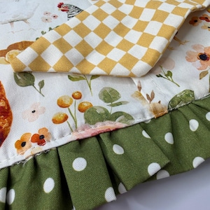 May include: Close-up of a fabric with a floral and chicken pattern. A yellow and white checkered fabric is on top. The bottom edge has a green polka dot ruffle.