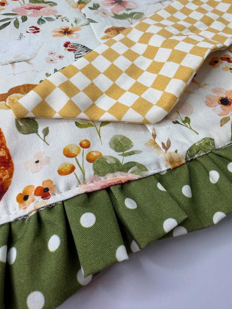 May include: Close-up of a fabric with a floral and chicken pattern. A yellow and white checkered fabric is on top. The bottom edge has a green polka dot ruffle.