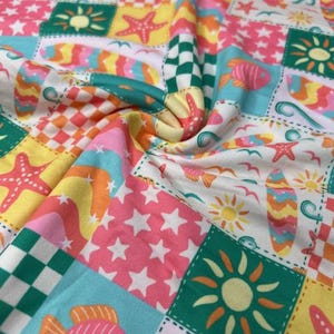 May include: A colorful fabric with a patchwork design featuring beach-themed illustrations. The pattern includes starfish, fish, surfboards, suns, and stars in a variety of colors, including pink, yellow, green, and blue. The fabric is folded, showing the detailed print.
