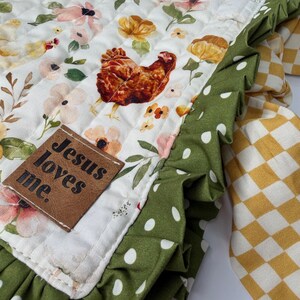 May include: A quilted fabric with a floral and chicken pattern. The main fabric is white with a repeating pattern of chickens, flowers, and foliage. A green polka dot fabric with a ruffled edge borders the main fabric. A brown leather tag reads "Jesus loves me."