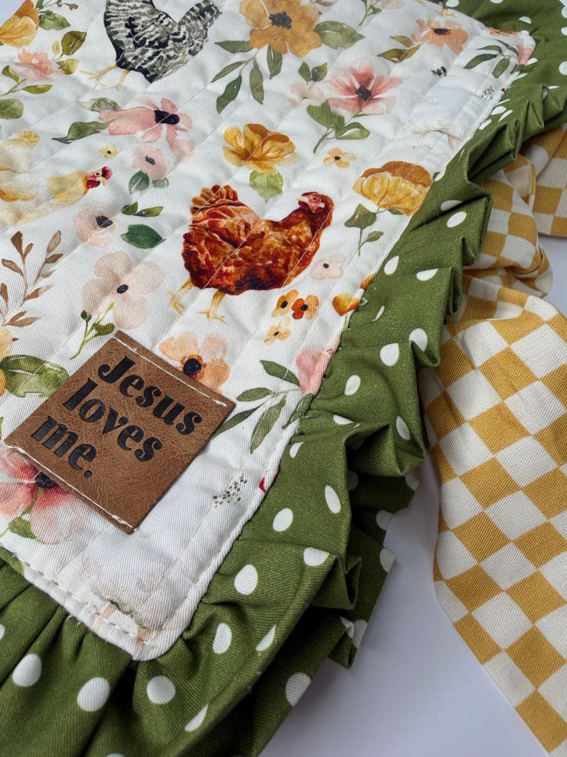 May include: A quilted fabric with a floral and chicken pattern. The main fabric is white with a repeating pattern of chickens, flowers, and foliage. A green polka dot fabric with a ruffled edge borders the main fabric. A brown leather tag reads "Jesus loves me."