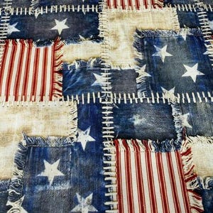 May include: A patchwork textile featuring a patriotic design. The design incorporates squares of faded blue denim with white stars, red and white striped fabric, and off-white fabric. The squares are stitched together with white thread.