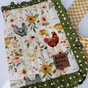 May include: A quilted blanket with a floral and chicken pattern. The blanket features a white background with pink, yellow, and orange flowers, and several chickens. The border is green with white polka dots, and a brown leather tag reads "Jesus loves me."