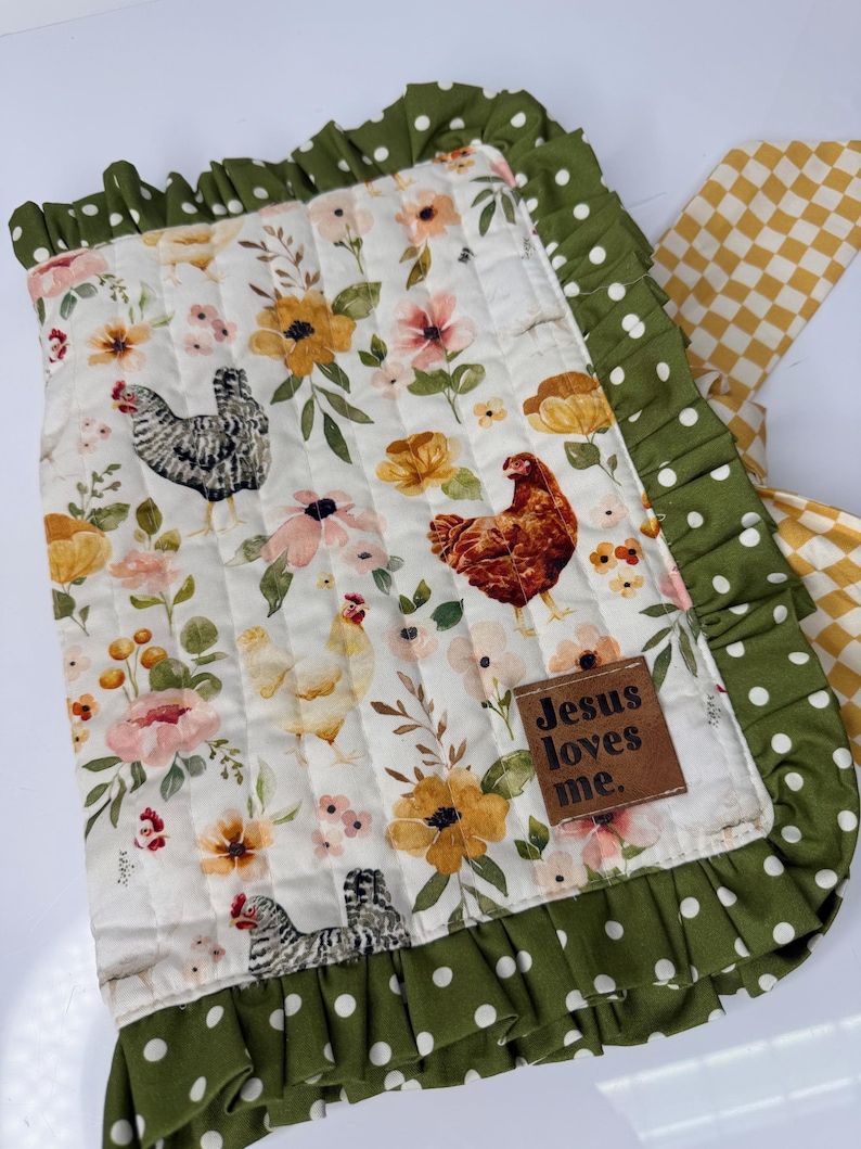 May include: A quilted blanket with a floral and chicken pattern. The blanket features a white background with pink, yellow, and orange flowers, and several chickens. The border is green with white polka dots, and a brown leather tag reads "Jesus loves me."