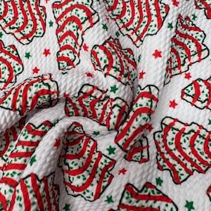 May include: White fabric with a repeating pattern of red and green Christmas tree shapes and red and green stars. The trees have red stripes and green dotted accents. The fabric has a textured surface.