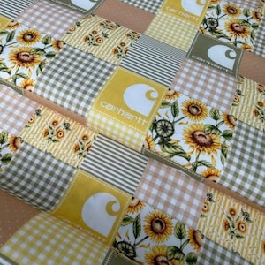 May include: A patchwork quilt featuring squares of various patterns and colors. The quilt includes squares with sunflowers, gingham, stripes, and Carhartt logos. The color palette consists of yellow, brown, and green.