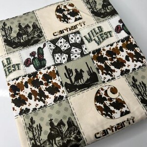 Op de afbeelding: Een patchwork stof met een witte achtergrond en verschillende patronen, waaronder een bruin en wit koeienprint, speelkaarten, cactussen en cowboys die op paarden rijden. De stof heeft ook het Carhartt-logo en de woorden "Wild West".
